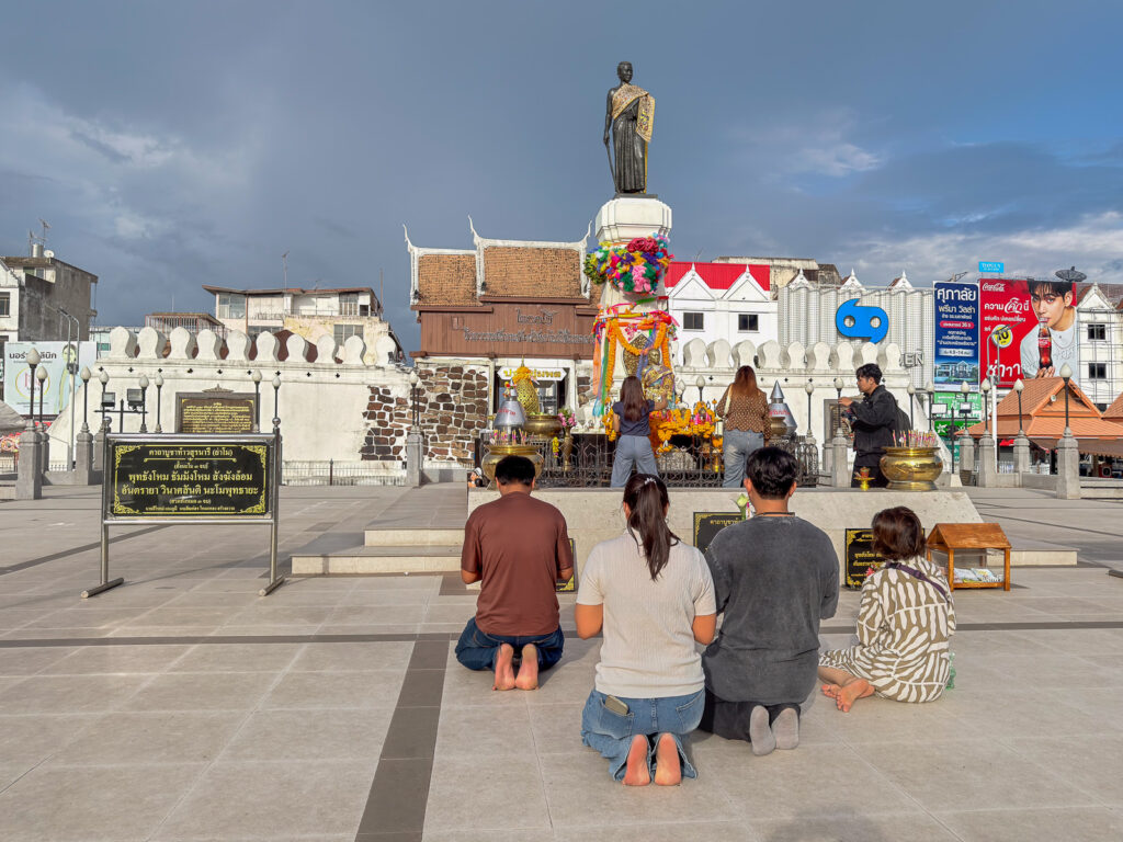 Personas arrodilladas ante el monumento a Thao Suranari, decorado con flores y ofrendas, junto a la puerta Chumphon en Korat.