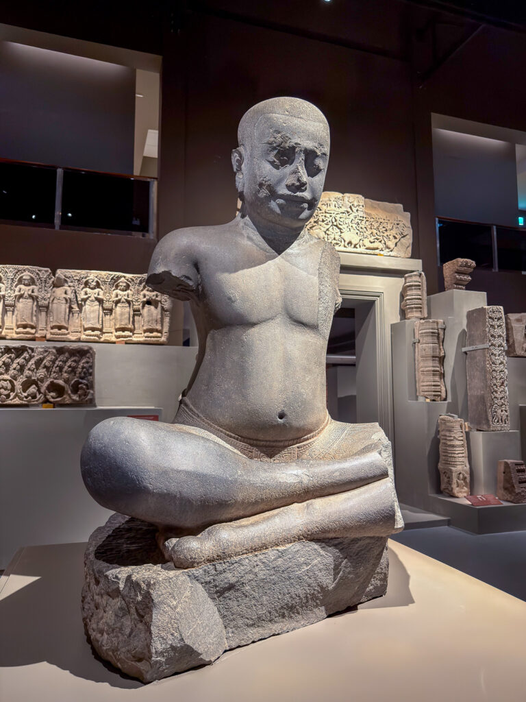 Escultura masculina de piedra sentada con las piernas cruzadas y los brazos incompletos