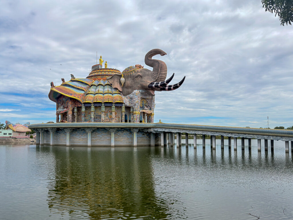 Pabellón con forma de elefante de Wat Ban Rai, decorado con mosaicos y reflejado en el estanque, junto a la pasarela de acceso.
